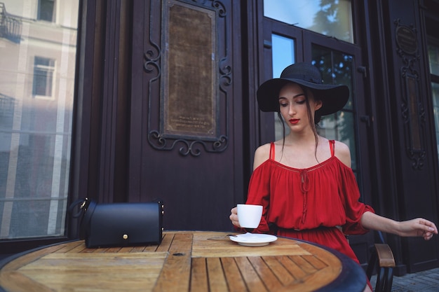 Junge elegante Frau in einem roten Kleid und in einem Weinlesehut mit einer Tasse Kaffee, die im Coffeeshop sitzt