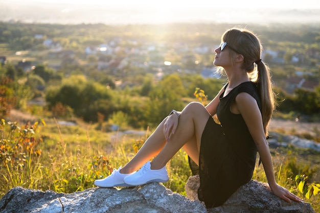 Junge elegante Frau in schwarzem kurzen Kleid und weißen Turnschuhen sitzt auf einem Felsen und entspannt sich am Sommerabend im Freien. Modische Dame, die warmen Sonnenuntergang in der Natur genießt.