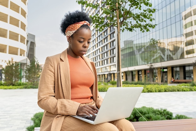 Junge elegante Geschäftsfrau afrikanischer Abstammung mit Laptop auf den Knien, die auf einer Holzbank im Park gegen Bürogebäude sitzt