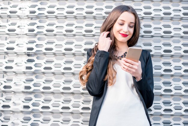 Junge elegante Geschäftsfrau im Anzug, die draußen mit Telefon nahe der modernen Metallwand steht