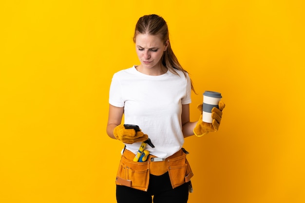 Junge Elektrikerin lokalisiert auf gelber Wand, die Kaffee hält, um und ein Handy wegzunehmen