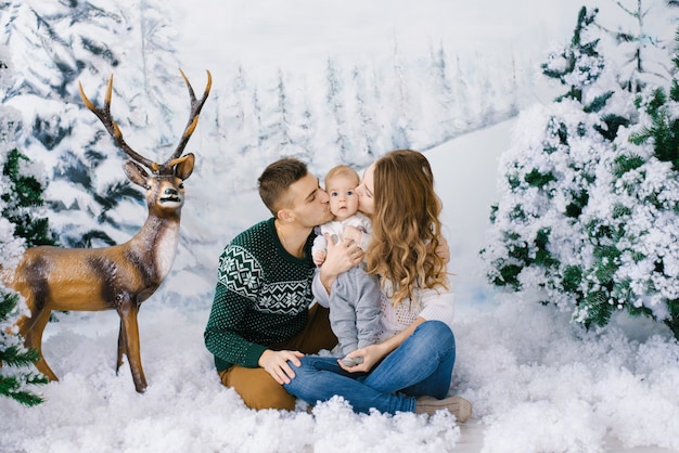 Junge eltern mit einem baby küssen das baby auf die wangen und sitzen im kunstschnee