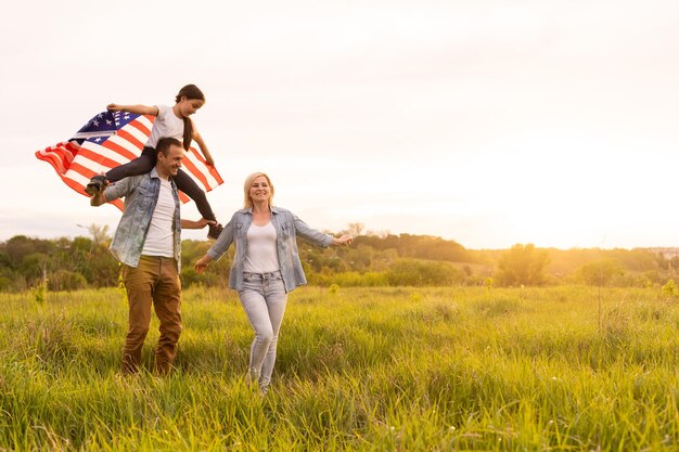 Junge Eltern mit ihrer Tochter, die bei Sonnenuntergang die amerikanische Flagge auf dem Land hält. Feier zum Unabhängigkeitstag.