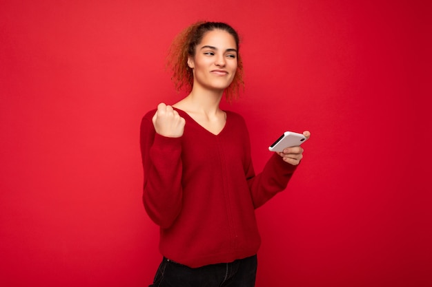 Junge emotionale Frau, die einen dunkelroten Pullover trägt, isoliert auf rotem Hintergrund, der ein Smartphone hält, das Spaß hat und eine Ja-Geste zeigt, die zur Seite schaut.