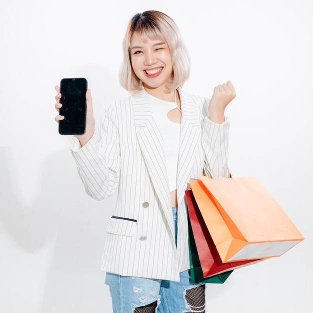 Junge energische asiatische Frau mit leeren Smartphone-Suche Shop-Einzelhandel mit bunten Einkaufstüten auf weißem Hintergrund Online-Shopping futuristisches Konzept