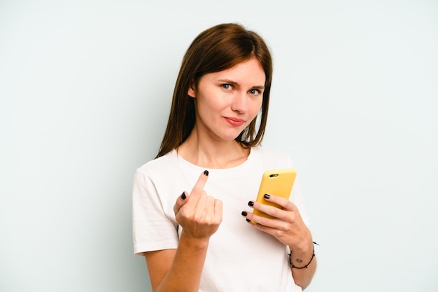 Junge Engländerin, die ein Handy isoliert auf blauem Hintergrund hält und mit dem Finger auf Sie zeigt, als würde sie einladen, näher zu kommen