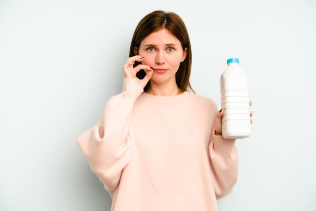 Junge Engländerin hält eine Flasche Milch isoliert auf blauem Hintergrund mit Fingern auf den Lippen, die ein Geheimnis bewahren.