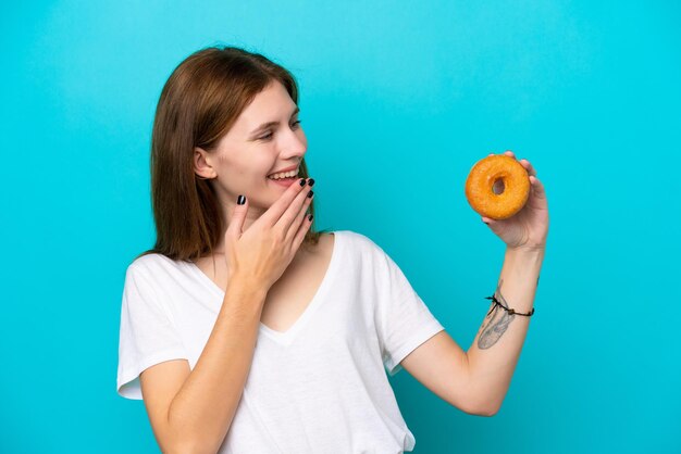Junge Engländerin hält einen Donut über isoliertem blauem Hintergrund mit überraschtem und schockiertem Gesichtsausdruck