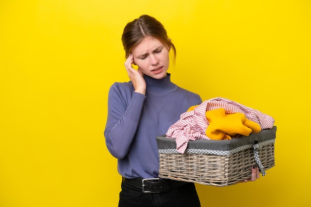 Junge Engländerin hält einen Kleiderkorb isoliert auf gelbem Hintergrund mit Kopfschmerzen