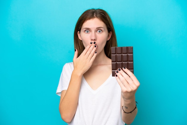 Junge Engländerin isoliert auf blauem Hintergrund, die eine Schokoladentafel nimmt und überrascht