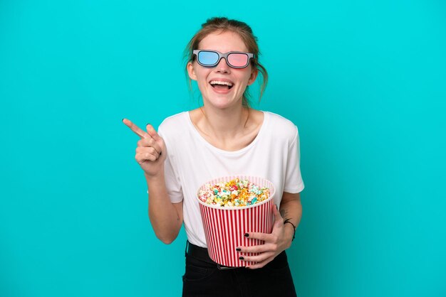 Junge Engländerin isoliert auf blauem Hintergrund mit 3D-Brille und hält einen großen Eimer Popcorn, während sie nach vorne zeigt