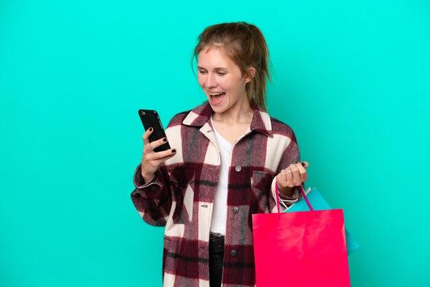 Junge Engländerin isoliert auf blauem Hintergrund mit Einkaufstüten und Schreiben einer Nachricht mit ihrem Handy an einen Freund
