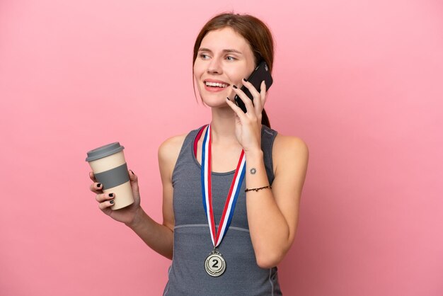 Junge Engländerin mit Medaillen isoliert auf rosa Hintergrund mit Kaffee zum Mitnehmen und einem Handy