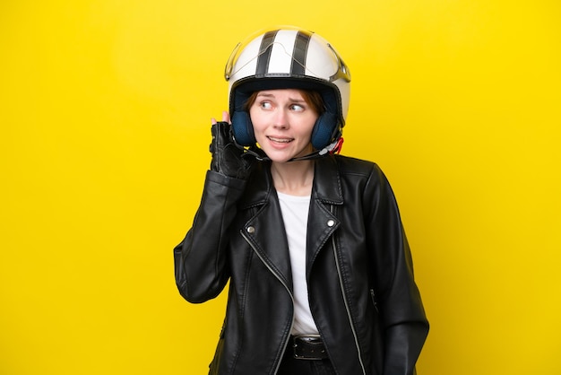 Junge Engländerin mit Motorradhelm isoliert auf gelbem Hintergrund, die etwas hört, indem sie die Hand auf das Ohr legt
