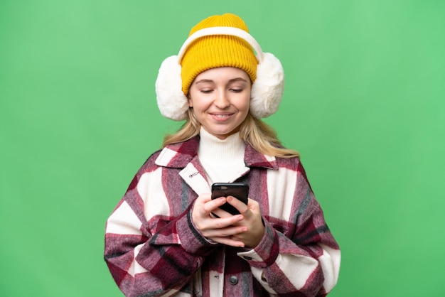 Junge Engländerin mit Wintermuffs vor isoliertem Hintergrund, die eine Nachricht mit dem Handy sendet