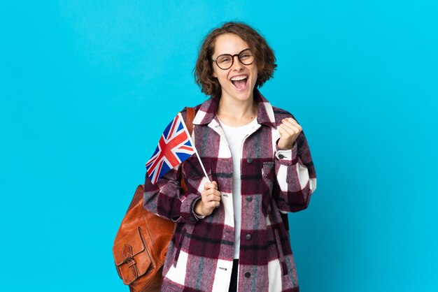 Junge englische Frau, die eine britische Flagge lokalisiert auf blauer Wand hält, die einen Sieg in der Siegerposition feiert