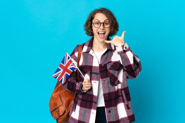 Junge englische Frau, die eine britische Flagge lokalisiert auf der blauen Wand hält, die Telefongeste macht. Rufen Sie mich zurück Zeichen