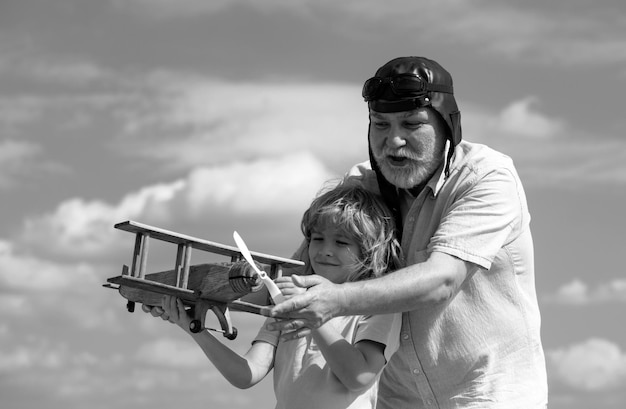 Junge Enkel und alter Großvater spielen mit einem Holzflugzeug gegen den Sommerhimmel-Hintergrund.