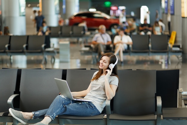 Junge entspannte Reisende Touristenfrau mit Kopfhörern, die Musik am Laptop hören, warten in der Lobbyhalle am internationalen Flughafen?