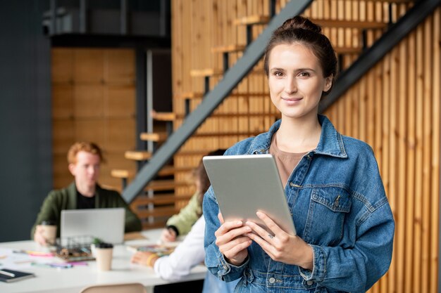 Junge erfolgreiche brünette Geschäftsfrau mit digitalem Tablet, die in der Arbeitsumgebung vor der Kamera steht und im Netz surft