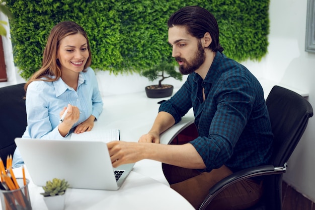 Junge erfolgreiche Designer sprechen im Büro über Projekt. Ein Mann spricht über ein Projekt, das auf einem Laptop ausgestellt ist, und eine junge Frau hört ihrer Kollegin zu und schaut auf den Bildschirm.