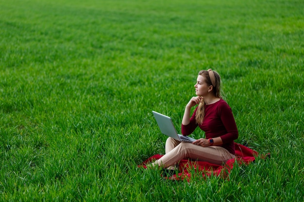 Junge erfolgreiche Frau sitzt auf grünem Gras mit einem Laptop in ihren Händen. Arbeite an der Natur. Studentenmädchen, das an einem abgelegenen Ort arbeitet. Neue Geschäftsideen