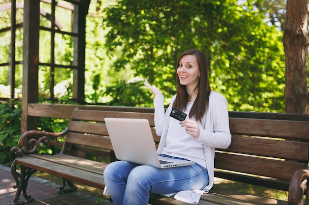 Junge erfolgreiche Geschäftsfrau, die Kreditkarte hält. Frau, die auf der Bank sitzt und an einem modernen Laptop-PC im Stadtpark in der Straße draußen auf der Natur arbeitet. Mobiles Büro. Freiberuflich, Geschäftskonzept.