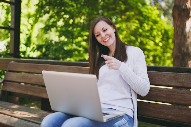 Junge erfolgreiche Geschäftsfrau in leichter Freizeitkleidung. Frau, die auf der Bank sitzt und an einem modernen Laptop-PC im Stadtpark in der Straße draußen auf der Natur arbeitet. Mobiles Büro. Freiberufliches Geschäftskonzept