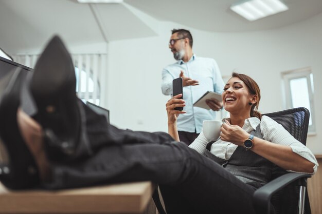 Junge erfolgreiche Unternehmerin schaut sich Smartphone an und trinkt Kaffee im Büro.