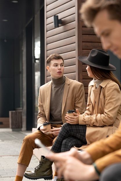 Junge erholsame Frau mit Getränk, die ihren Freund anschaut, während sie beide auf der Bank von moderner Architektur sitzt und sich beim Chillen oder Dating unterhalten?