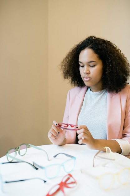 Junge ernste Frau in der intelligenten lässigen Betrachtung einer Brille in ihren Händen mit einer Vielzahl von Brillen auf dem Tisch