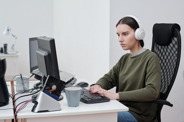Junge ernsthafte Frau in Freizeitkleidung, die bei der Arbeit Musik über Kopfhörer hört