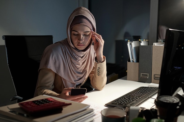 Junge ernsthafte Geschäftsfrau im Hijab, die spät in der Nacht im Smartphone scrollt