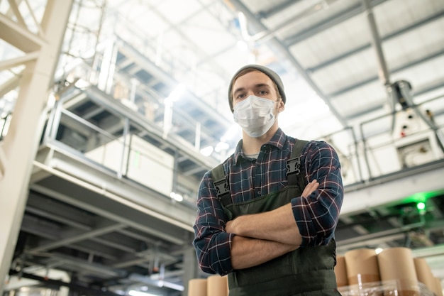 Junge ernsthafte männliche Arbeiter einer großen Fabrik in Arbeitskleidung und Schutzmaske, die mit verschränkten Armen vor der Kamera im Lager steht