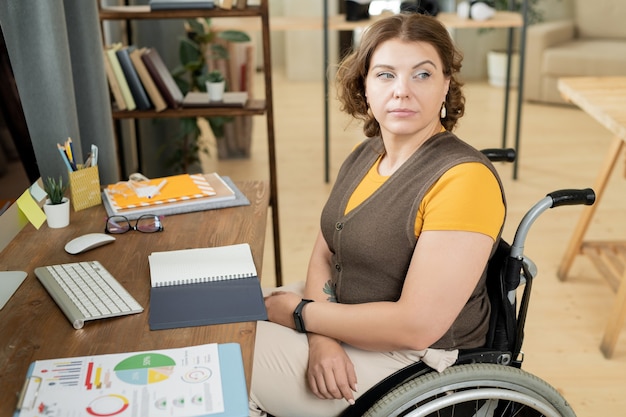 Junge ernsthafte oder nachdenkliche Geschäftsfrau im Rollstuhl, die beiseite schaut, während sie am Schreibtisch vor dem Computer sitzt und mit Daten im Büro arbeitet