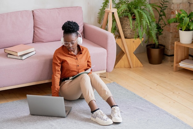 Junge ernsthafte schwarze Frau in Freizeitkleidung und Kopfhörern, die während des Online-Unterrichts auf dem Boden neben der Couch vor dem Laptop sitzen