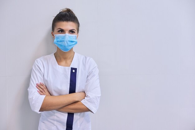 Junge ernsthafte selbstbewusste Ärztin in medizinischer Maske mit verschränkten Armen, Hintergrundwand zum Krankenhaus, Kopienraum