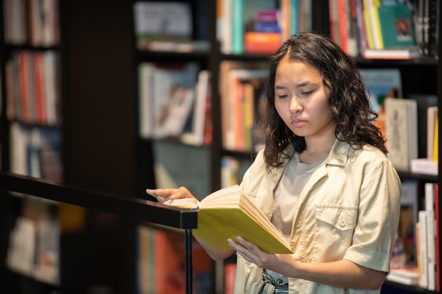 Junge ernsthafte Studentin liest ein Buch, während sie am großen Bücherregal steht