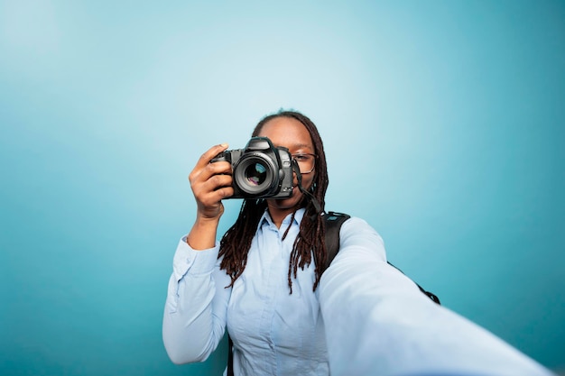 Junge erwachsene afrikanisch-amerikanische Frau, die ein DSLR-Gerät auf die Kamera zeigt, während sie ein Foto auf blauem Hintergrund macht. Kreativer Amateurfotograf, der Bilder mit moderner Kamera für soziale Medien macht.
