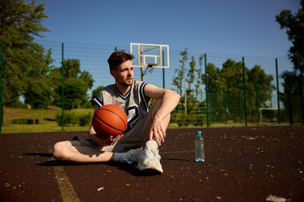 Junge erwachsene Basketballspieler ruhen sich auf einem städtischen Straßenplatz aus. Sportler in Mannschaftsuniform hält den Ball und entspannt sich nach dem Spiel