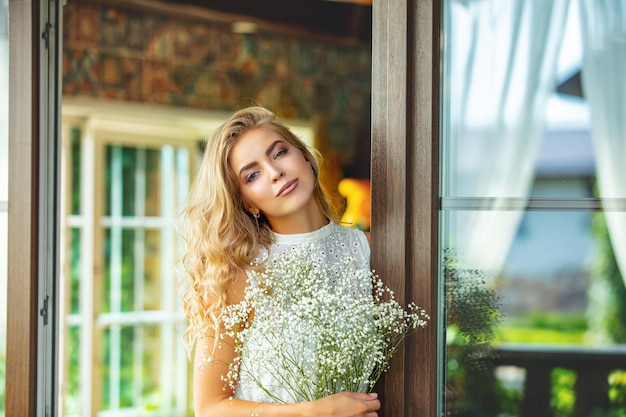 Foto junge erwachsene blonde schönheit in einem weißen kleid an den fenstern auf dem balkon mit weißen blumen