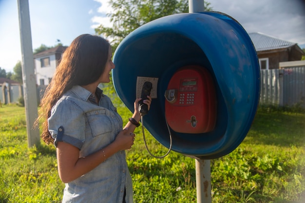 Junge erwachsene Frau, die an einem öffentlichen Straßentelefon auf der Straße in einem russischen Dorf bei Sonnenschein spricht