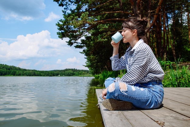 Junge erwachsene Frau, die auf einem Holzsteg sitzt, Kaffee trinkt und auf den See blickt