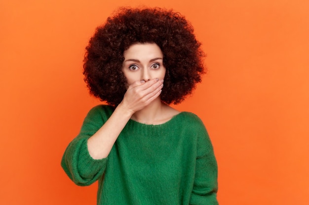 Junge erwachsene Frau mit Afro-Frisur, die einen grünen Pullover im lässigen Stil trägt, der den Mund mit einer Handfläche bedeckt, geheim halten, sagt es niemandem Indoor-Studioaufnahme isoliert auf orangefarbenem Hintergrund