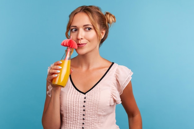 Junge erwachsene Frau mit blonden Haaren im Sommerkleid, die frische Cocktails trinkt und mit verträumtem Gesichtsausdruck wegschaut und tropischen Urlaub genießt Studioaufnahme isoliert auf blauem Hintergrund