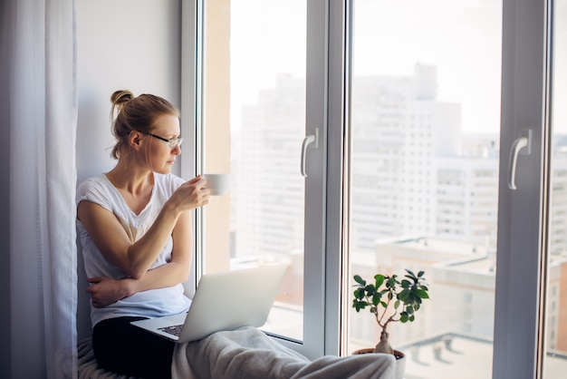 Junge erwachsene Frau mit Brille und weißem T-Shirt sitzt auf der Fensterbank mit Laptop zu Hause, trinkt Kaffee und schaut auf das Fenster. Remote-Arbeit, freiberuflich, Quarantäne, Selbstisolation, Sperrung.