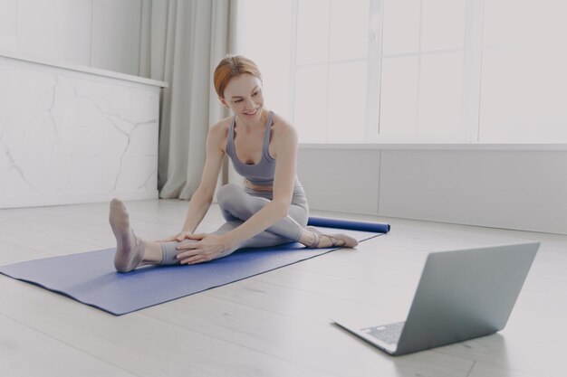 Junge erwachsene glückliche Frau, die Yoga beim Stretching von Asanas durch Online-Unterricht genießt