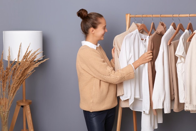 Junge erwachsene glückliche Frau mit Brötchenfrisur, professionelle Modestylistin, die in der Nähe von Kleidern steht, hängt im Regal im Designergeschäft, das trendiges Outfit in der Shop-Boutique verkauft