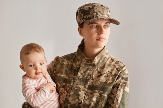 Junge erwachsene kaukasische Soldatin mit ihrer kleinen süßen Tochter in den Händen, die mit ernstem traurigem Gesichtsausdruck wegschaut und nach Krieg oder Armee nach Hause zurückkehrt.