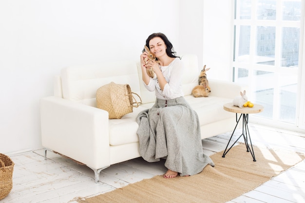 Junge erwachsene schöne Frau mit flauschigen Kaninchen und Ostereiern, die auf einem weißen Sofa sitzen